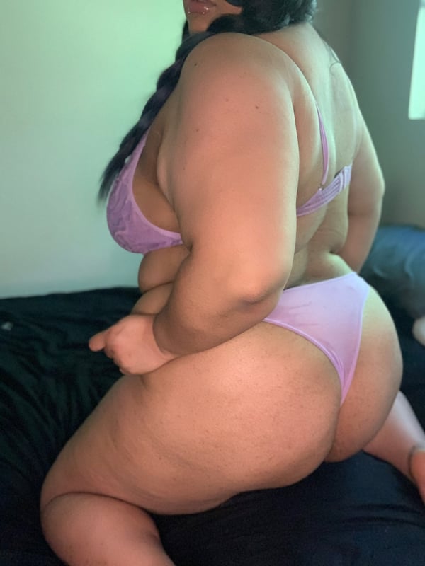 A curvy woman with braided hair sitting on a bed, wearing a purple swimsuit and posing confidently indoors.