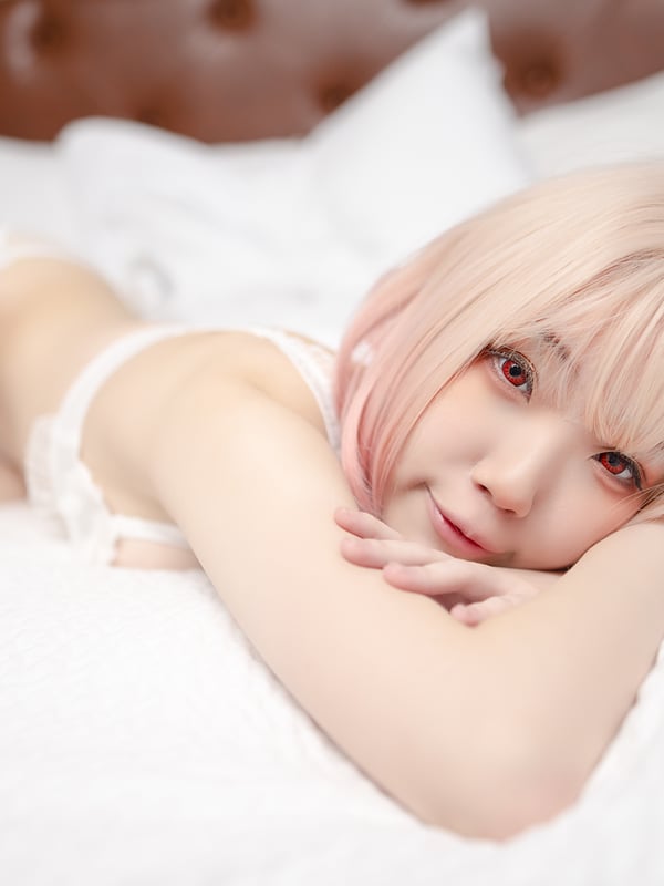 Young woman with pink hair lying on a bed, wearing white lingerie, smiling softly against white pillows and a wooden headboard.