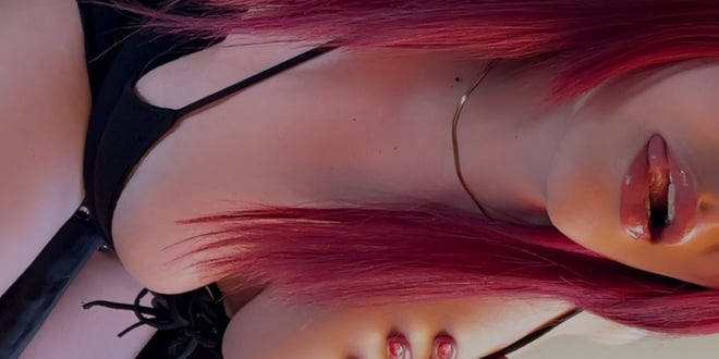 Close-up portrait of a woman with long pink hair, wearing a black top and gold necklace, posing confidently with glossy lips.