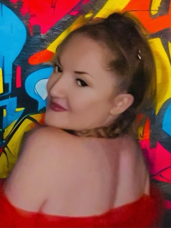 A woman with brunette hair in a ponytail smiling over her shoulder against a vibrant graffiti wall, dressed in a red sheer top.