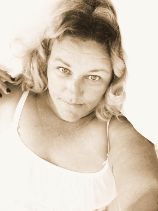 A woman with blonde curly hair posing confidently in a white tank top against a light background.