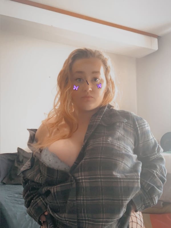 A woman with blonde hair and sparkly eye effects smiling in an open plaid shirt and fishnet stockings indoors.