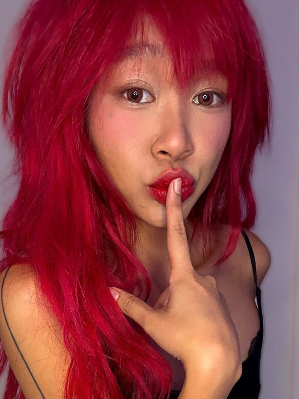 A young woman with bright red hair making a playful shushing gesture with her finger over her lips, wearing a black top.