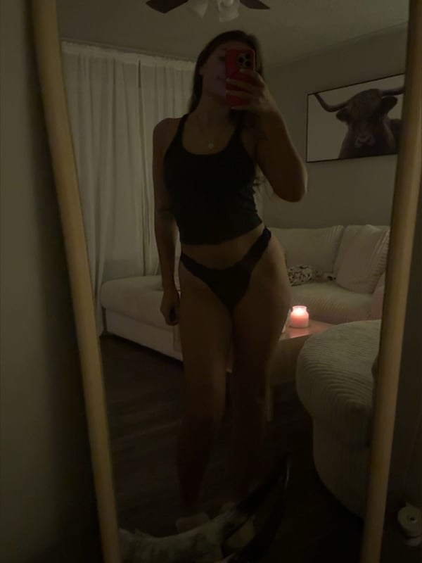 A woman taking a mirror selfie in a tank top and underwear inside a room with a ceiling fan and couch.