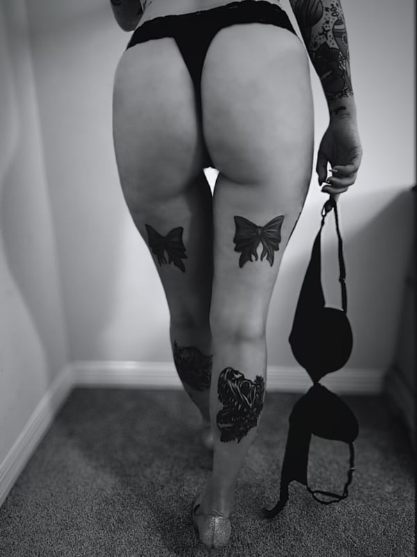 A woman with tattoos stands facing away, wearing a thong and high heels while holding a bra, viewed from behind in a room.