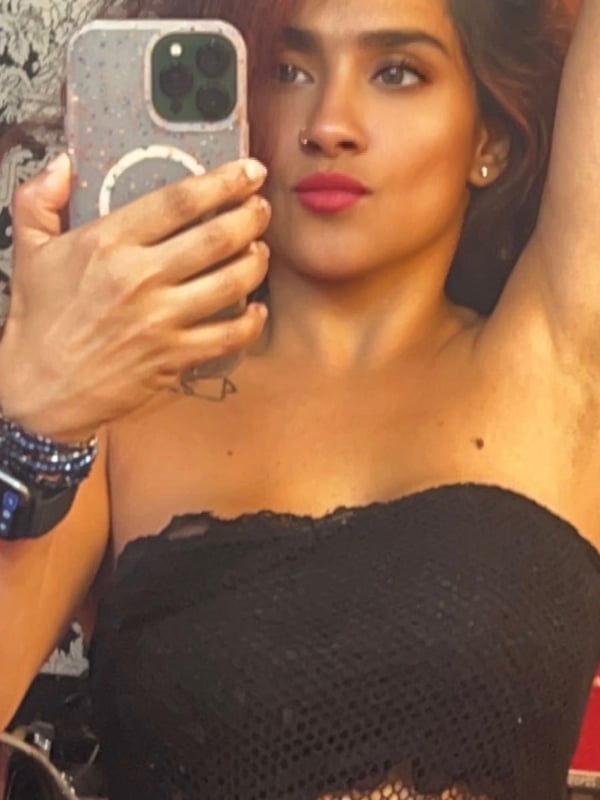 A young woman taking a selfie in a black lace top, holding her phone with one hand raised, accessorized with bracelets and earrings against a decorative background.