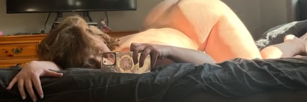 A person lying on a bed in a cozy bedroom with warm lighting, wearing orange clothing and glasses, surrounded by furniture.