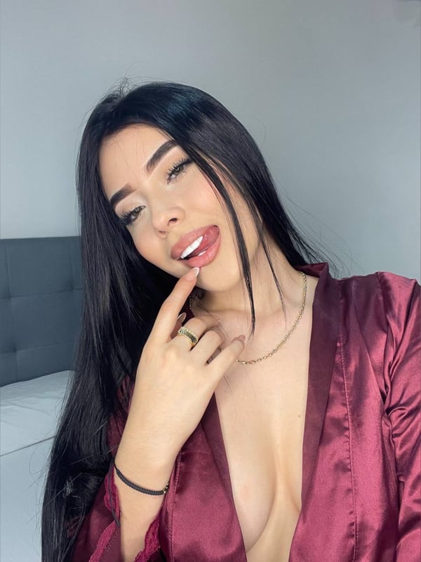 Close-up selfie of a young woman with long dark hair, wearing a maroon satin robe, smiling playfully against a neutral background.