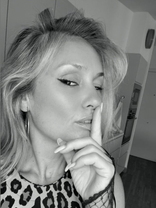 A woman with blonde hair making a shushing gesture with her finger over her lips, wearing a leopard print top with lace sleeves, in a black and white indoor setting.