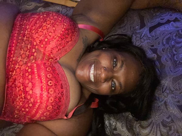 A woman smiling while lying on a bed, wearing a red lace top and posing relaxed against decorative pillows.