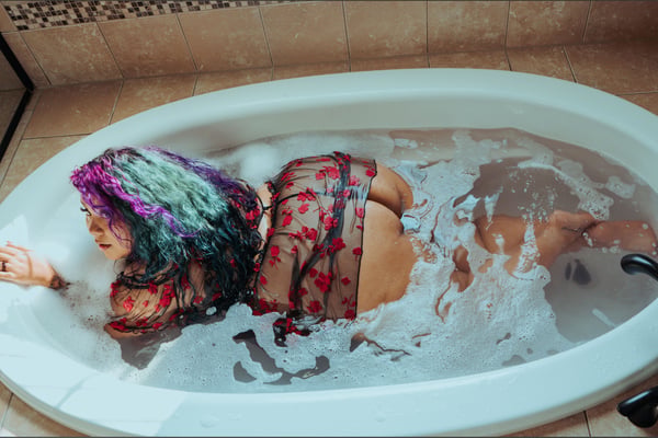A person with colorful hair reclining in a bathtub filled with bubbles, wearing a sheer red top, in a tiled bathroom setting.
