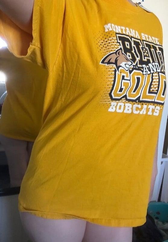 A woman wearing a yellow Montana State Bobcats t-shirt, lifting it slightly while posing indoors.