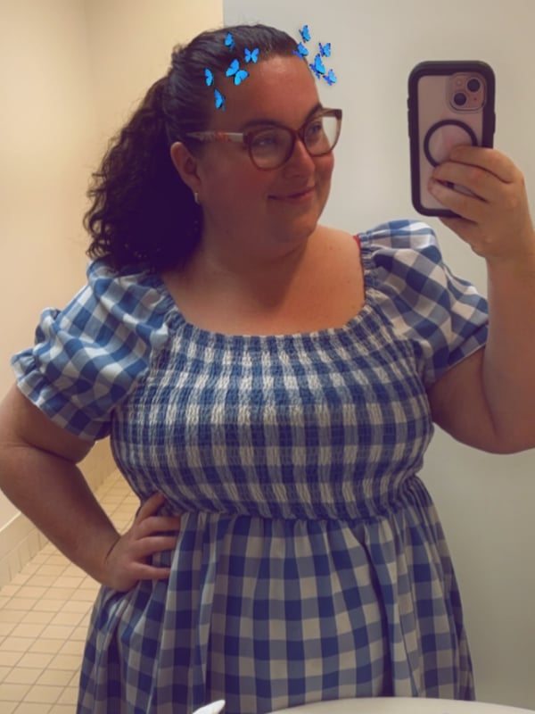 A woman with curly hair and blue hair clips smiling in glasses and a checkered dress, taking a mirror selfie in a bathroom.