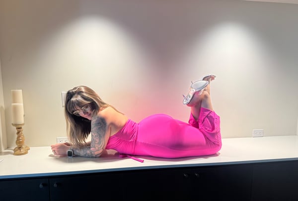 Woman lying on a white surface in a bright pink outfit, posing with legs raised near a lit candle.