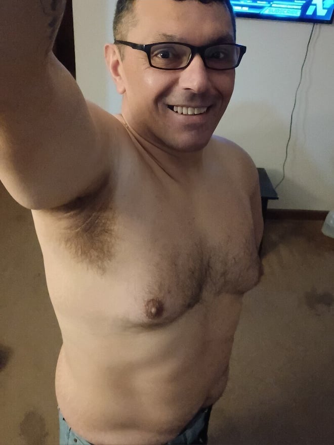 Shirtless man taking a selfie indoors, smiling with glasses, short dark hair, and visible armpit and chest hair.