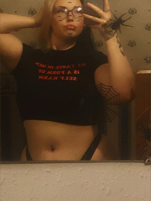 A young woman taking a mirror selfie in a bathroom with spider web decorations, wearing glasses, a graphic t-shirt, and bracelets.