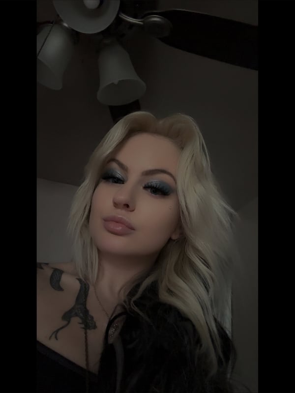 Close-up portrait of a woman with long blonde hair, dramatic eye makeup, and tattoos visible on her shoulder, indoors with a ceiling fan overhead.