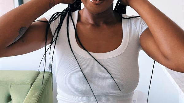 A woman with braided hair wearing a white top, posing with hands near her face against a green background.