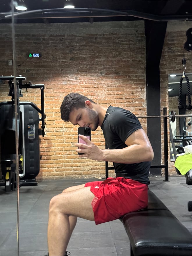 A man taking a selfie in a black t-shirt and red shorts while seated on a gym bench against a brick wall with exercise equipment nearby.