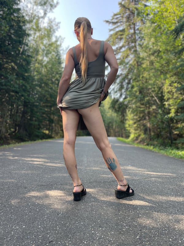 Woman with blonde ponytail standing on a forest road, wearing a short green dress and sandals, viewed from behind.