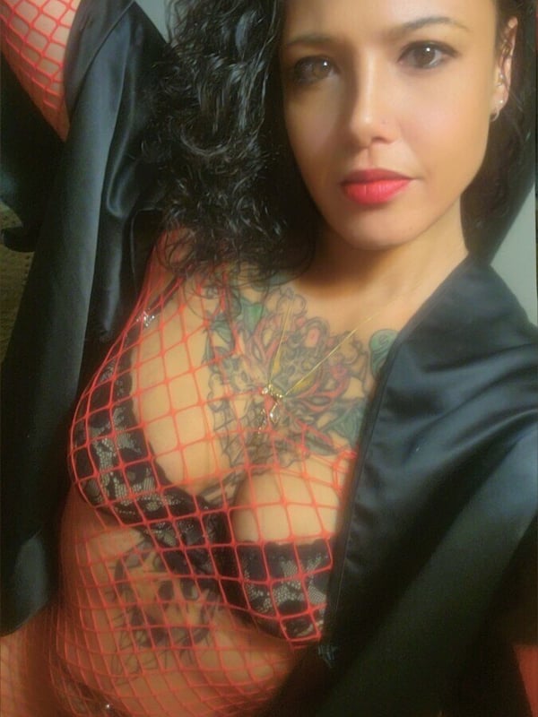A woman with tattoos and curly hair stands confidently in a black sheer top, smiling at the camera indoors.