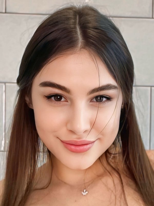 A young woman with long brown hair and subtle makeup smiles at the camera against a tiled wall.