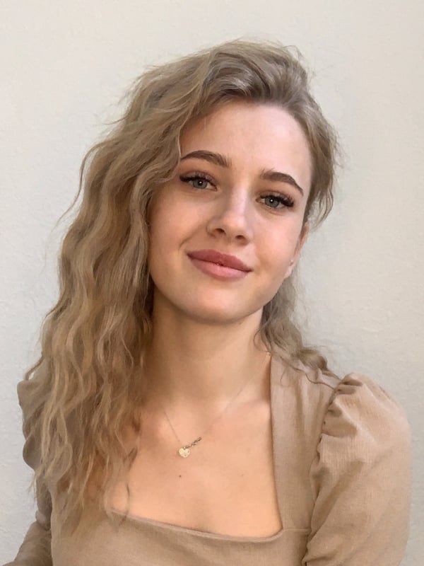 A young blonde woman with wavy hair smiling softly at the camera, wearing a light beige off-shoulder top and a delicate necklace against a plain background.