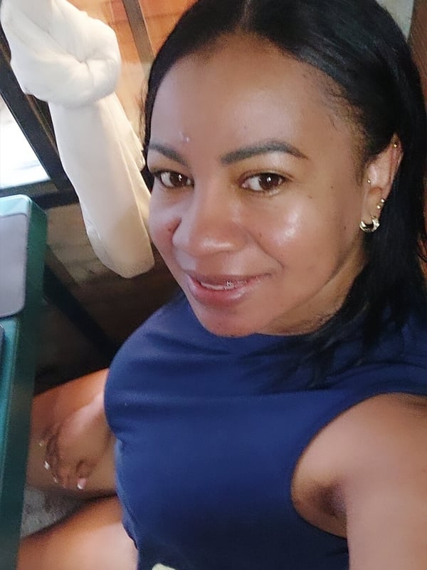 Woman with straight black hair smiling in a close-up selfie, wearing a blue sleeveless top, posed against a wooden background with white fabric.