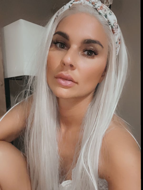 A woman with long white hair wearing a floral headband, posing indoors near a lamp with a neutral background.