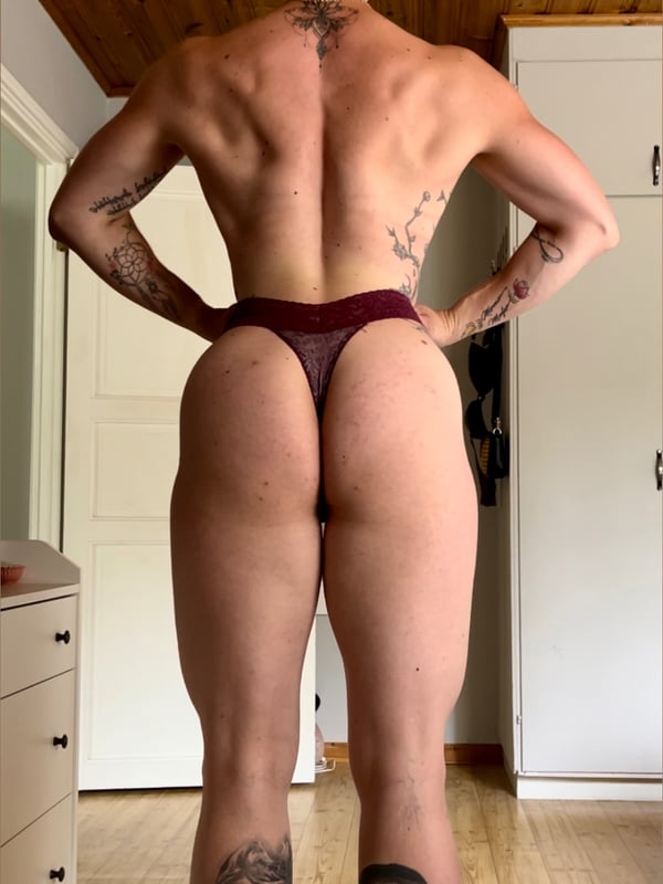 A muscular person posing shirtless from behind in underwear inside a room with wooden ceiling and furniture.