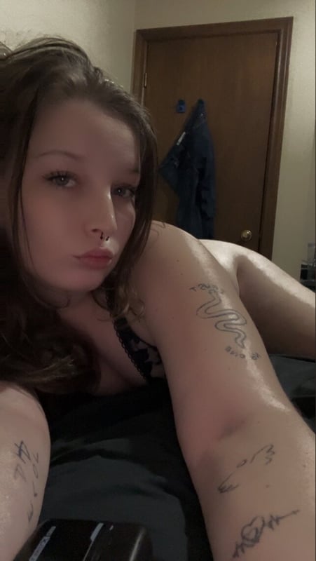 Young woman with long brown hair lying on a bed in lingerie, posing confidently in a cozy bedroom with wooden door and hanging clothes.