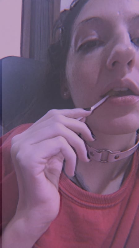 A young woman with dark hair wearing headphones and a choker necklace, posing close-up in a dimly lit indoor setting.