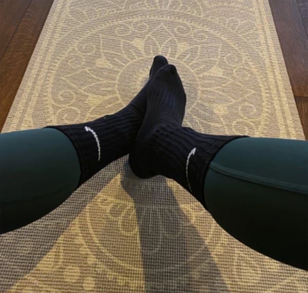 A person wearing black Nike socks and green leggings poses with legs extended on a patterned rug in a cozy indoor setting.