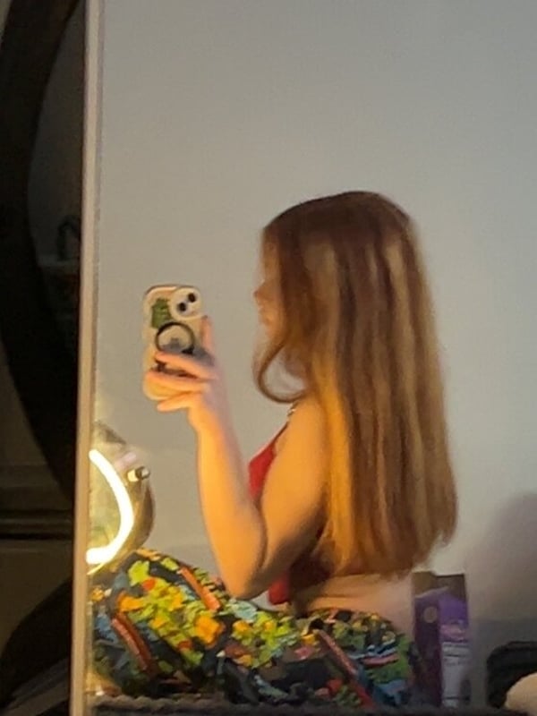 Young woman with long red hair taking a mirror selfie in a tank top and patterned pants, holding her phone in a bedroom setting.