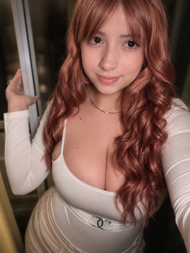 A close-up selfie of a young woman with long brown hair and bangs wearing a pink lace top.