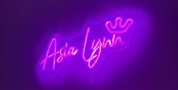 Neon pink glowing sign displaying the name 'Asia Lynnx' with a crown on a purple background.