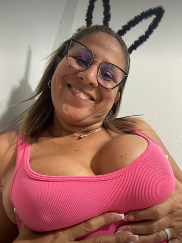 A smiling woman with bunny ears and glasses poses in a pink top, hands on chest against a plain background.