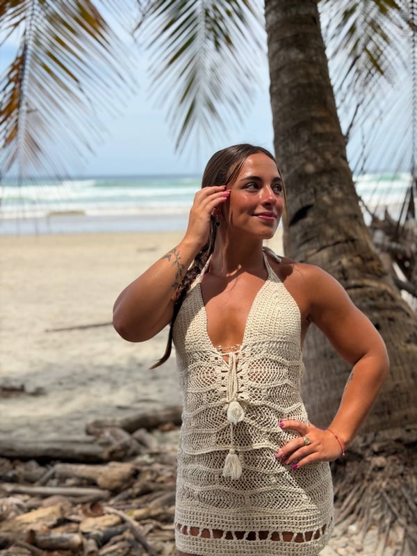 Athletic woman smiling and posing playfully on a sunny beach beside a palm tree, dressed in a crocheted top.