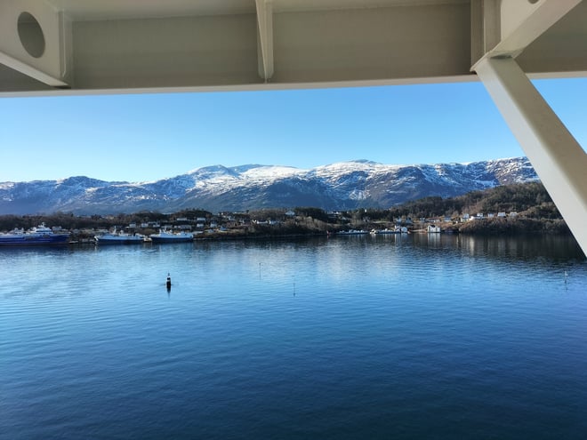 Scenic view of snow-capped mountains and a calm fjord with boats from a ship's deck on a clear blue day.