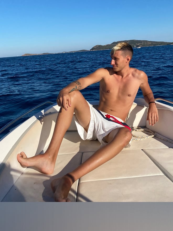 A shirtless man with tattoos sits on a boat deck, relaxing against the railing with a scenic sea and island view in the background.