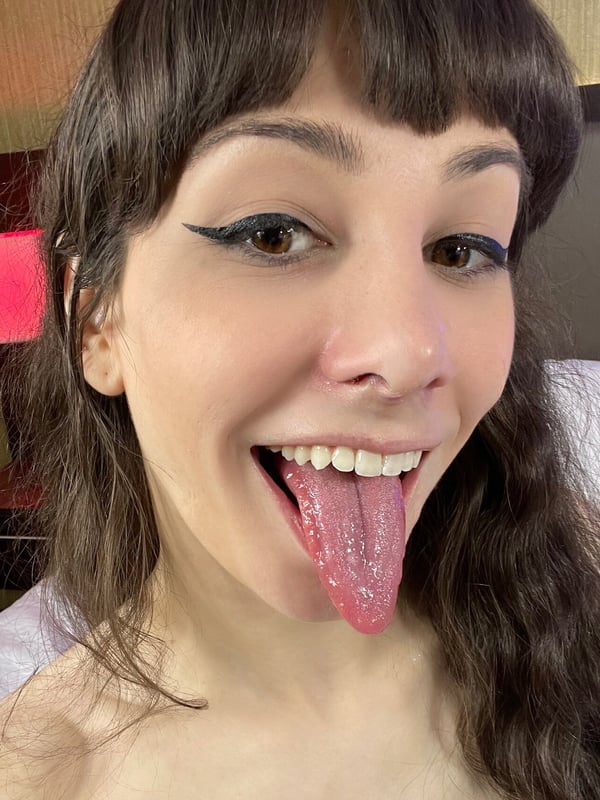 Close-up of a smiling young woman with bangs and eyeliner, sticking out her tongue in a playful expression against a bedroom background.