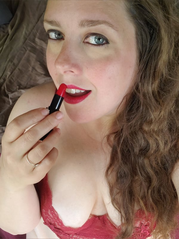 A woman with wavy brown hair applying red lipstick, wearing a red lace top while lying down.