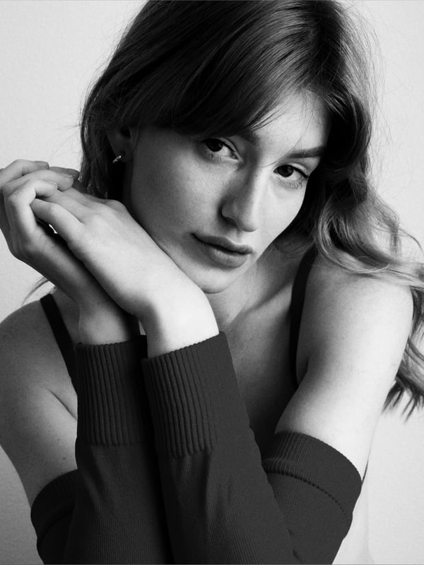 A woman with long wavy hair posing thoughtfully with hands near her face, wearing an off-shoulder top in a studio portrait.