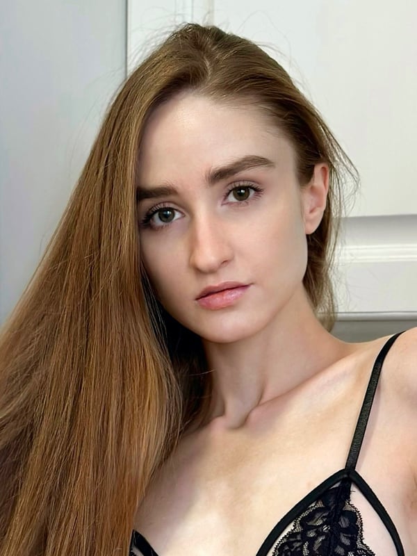 A young woman with long auburn hair and green eyes takes a close-up selfie, wearing a black lace bralette top against a light indoor background.