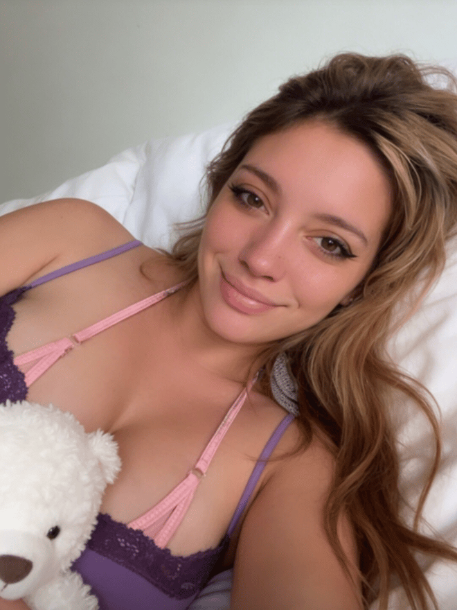 A young woman taking a selfie while lying down, wearing a purple and pink lace top and holding a white teddy bear.