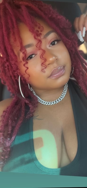 A woman with long red curly hair, wearing hoop earrings and a chain necklace, posing confidently in a car with a low-cut top.