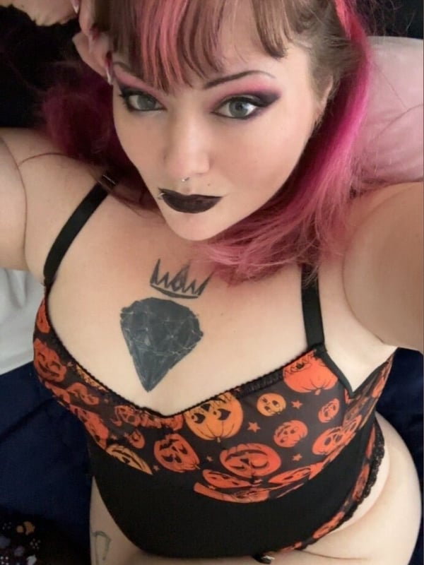 A woman with dramatic pink hair, heavy makeup, and a crown tattoo takes a close-up selfie in an orange Halloween-themed top.