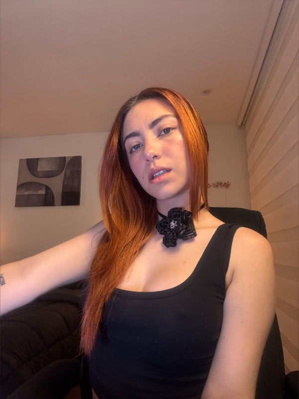A woman with long reddish hair wearing a black top and choker necklace, sitting indoors with a neutral expression.
