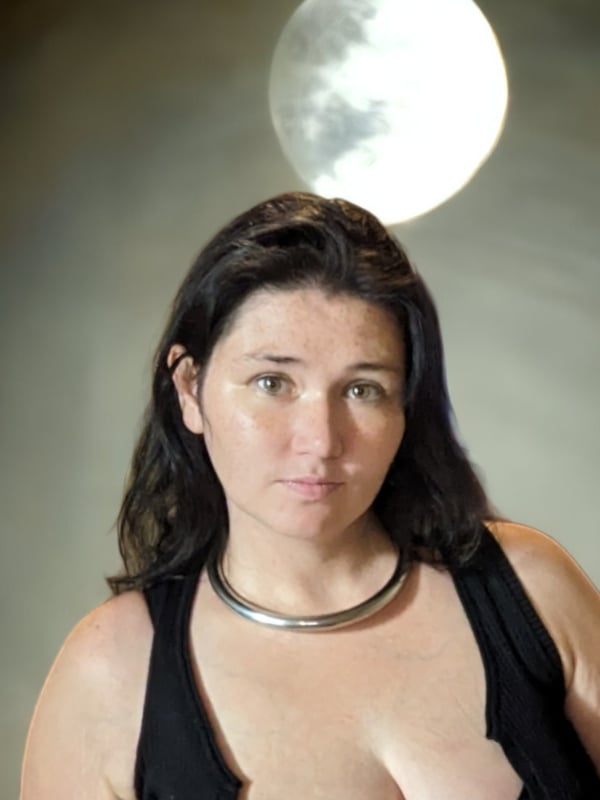 Close-up portrait of a woman with shoulder-length dark hair, wearing a black sleeveless top and choker necklace, standing against a dark background with a glowing full moon overhead.