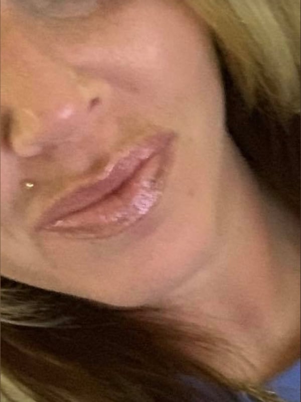Close-up selfie of a young woman with blonde hair, glossy lips, and a lip piercing, smiling softly while wearing a gold chain necklace.